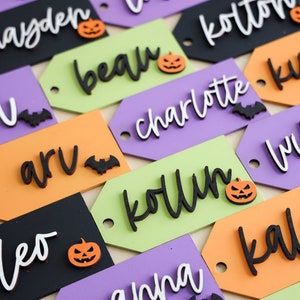 May include: A collection of Halloween-themed gift tags with various names written in black, white, and orange lettering. Each tag features a small pumpkin or bat decoration.