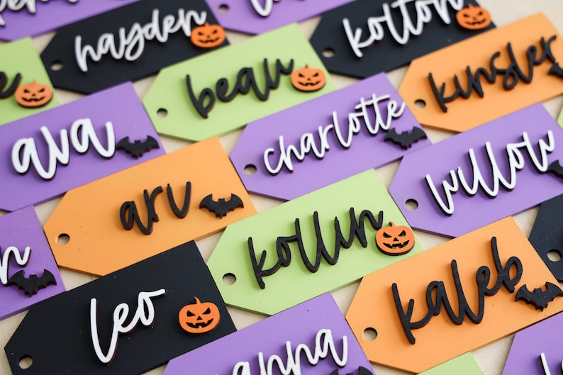 May include: A collection of Halloween-themed gift tags with various names written in black, white, and orange lettering. Each tag features a small pumpkin or bat decoration.