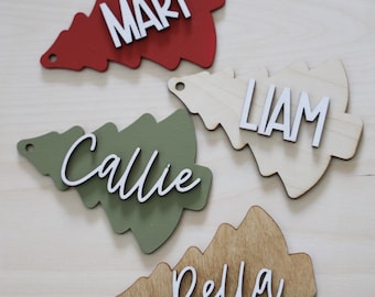 Könnte beinhalten: Vier Weihnachtsbaumschmuck aus Holz in Tannenbaumform, jeweils mit einem weißen Namen: "Mary" auf Rot, "Callie" auf Grün, "Liam" auf Naturholz und "Bella" auf Braun. Jedes hat ein Loch zum Aufhängen.