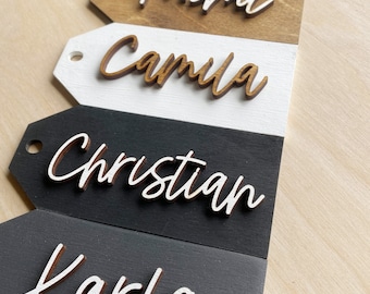 May include: Five wooden tags with names written in white script. The names are "Mommy", "Mimi", "Camila", "Christian", and "Karla". The tags are arranged vertically, with the top tag being "Mommy" and the bottom tag being "Karla".