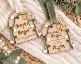 May include: Two arched wooden ornaments featuring the text Married + Bright, two gold-coloured rings, and a green vine with white light bulbs. One ornament displays The Muehlbergers 2024, the other First Christmas as Mr. & Mrs. Pecora 2024.