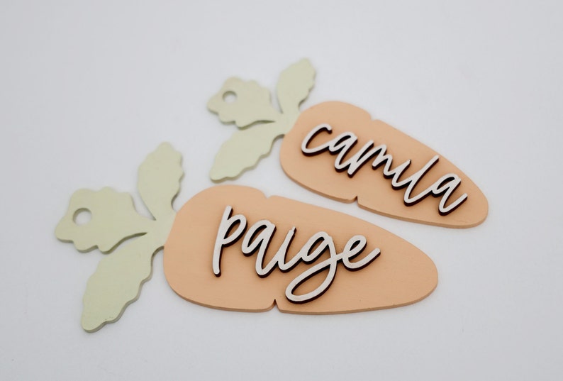 May include: Two wooden carrot-shaped ornaments with green leafy tops. The carrots are painted a light peach color and have the names "paige" and "camilla" written on them in a cursive font.