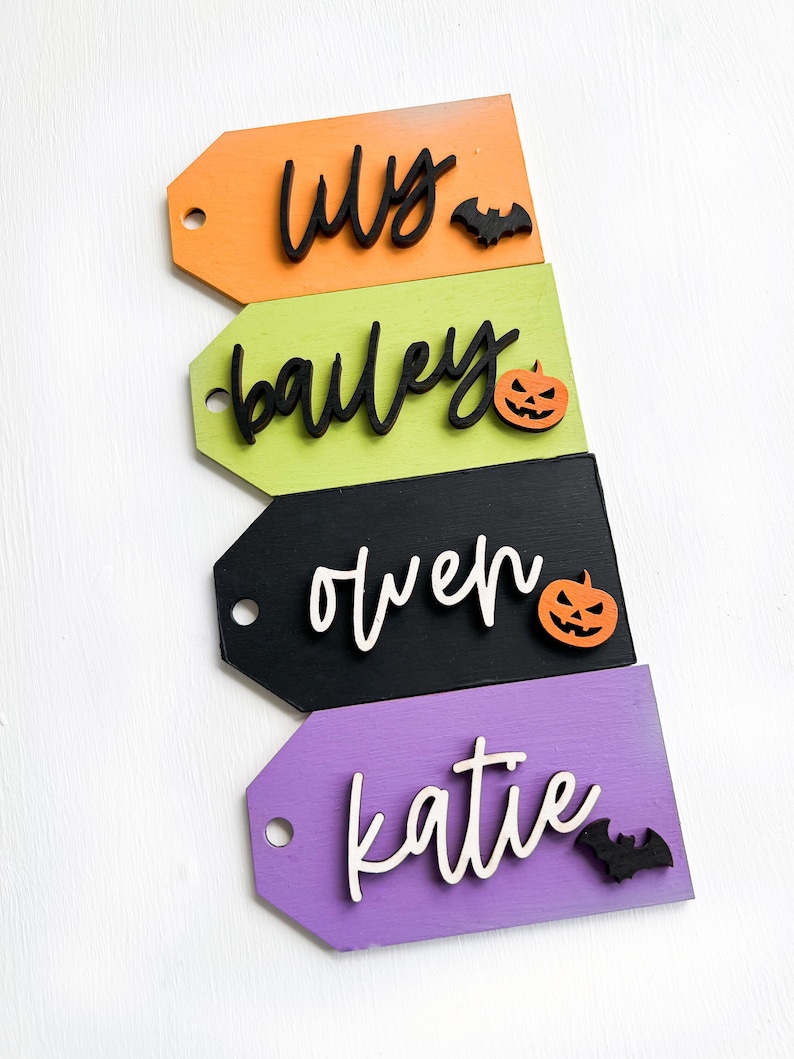 May include: Four wooden Halloween-themed gift tags with names written in black script. The tags are painted in orange, green, black, and purple. Each tag has a small wooden decoration, either a bat or a pumpkin.