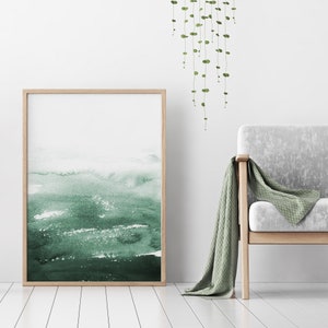 May include: A framed watercolour painting with a green and white abstract design. The painting is hanging on a white wall above a light grey armchair with a green throw blanket.