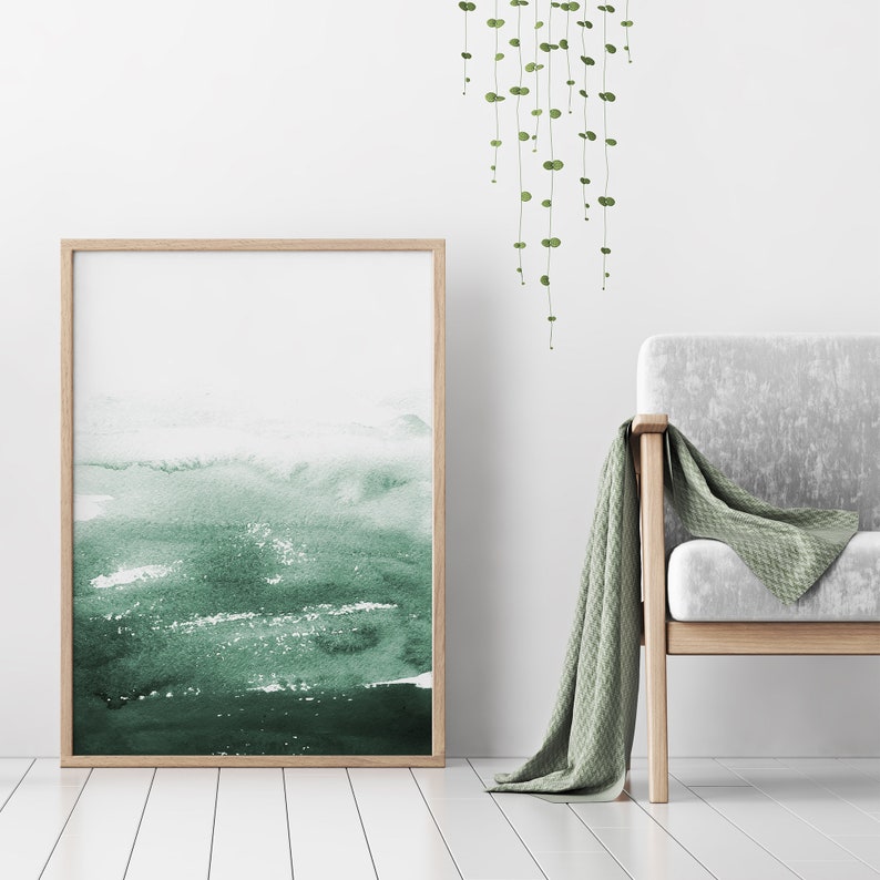 May include: A framed watercolour painting with a green and white abstract design. The painting is hanging on a white wall above a light grey armchair with a green throw blanket.