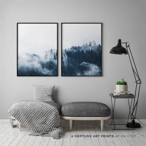 May include: Two framed prints of a misty forest scene in shades of blue. The prints are hung on a gray wall above a gray ottoman with a striped throw blanket.