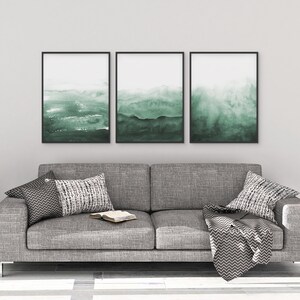 May include: Three framed abstract watercolour paintings in shades of green, hanging on a white wall above a grey sofa with cushions and a throw blanket.