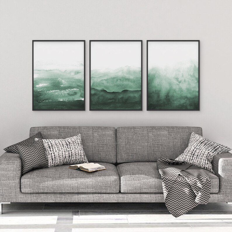 May include: Three framed abstract watercolour paintings in shades of green, hanging on a white wall above a grey sofa with cushions and a throw blanket.