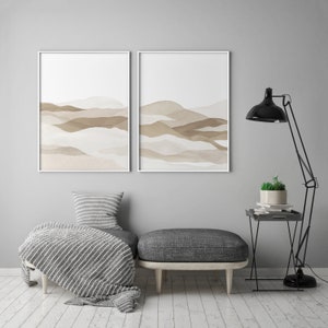 May include: Two framed abstract landscape prints with brown and beige watercolor mountains on a white background. The prints are hanging on a light gray wall above a gray ottoman with a striped throw blanket. A black floor lamp with a black shade is in the corner of the room.