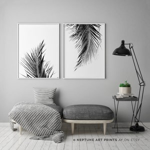 May include: Two black and white prints of palm leaves in simple white frames, hanging on a grey wall above a grey sofa with a striped throw blanket and a small side table with a potted plant.