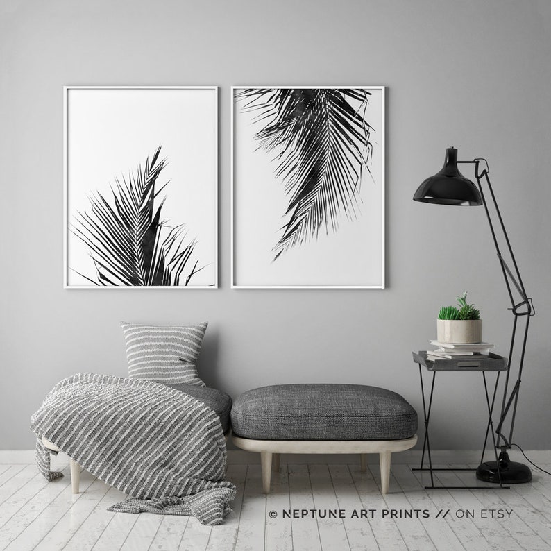 May include: Two black and white prints of palm leaves in simple white frames, hanging on a grey wall above a grey sofa with a striped throw blanket and a small side table with a potted plant.
