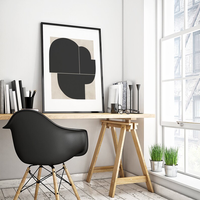 May include: A black and white abstract art print in a black frame hangs above a wooden desk with a black chair and a wooden sawhorse table. There are two potted plants on the windowsill.