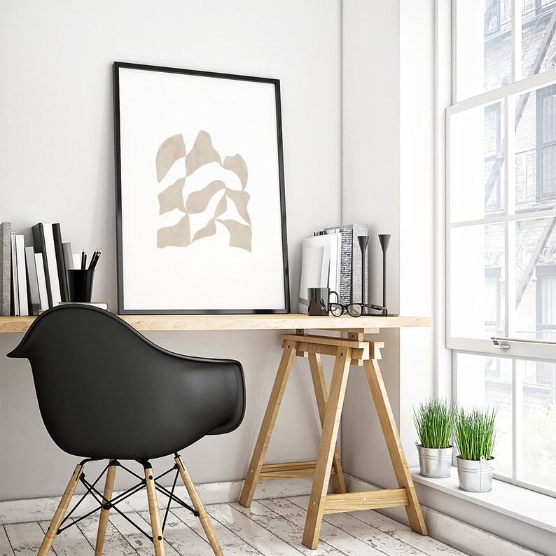 May include: A minimalist home office with a black chair, wooden desk, and a framed abstract print with beige shapes on a white background. The desk is on wooden sawhorses and there are two potted plants in the corner.