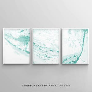 May include: Set of three abstract art prints featuring a white background with teal blue marbling. The prints are framed in white.