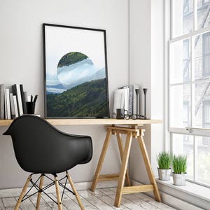 Peut inclure: Une impression encadr&eacute;e noire d'un paysage avec une d&eacute;coupe circulaire r&eacute;v&eacute;lant une cha&icirc;ne de montagnes et des nuages. L'impression est accroch&eacute;e &agrave; un mur blanc au-dessus d'un bureau en bois avec une chaise noire, un chevalet en bois et deux plantes en pot.