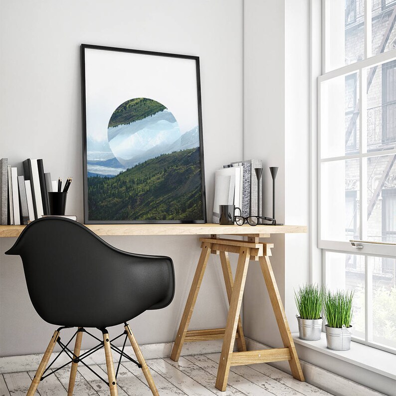 Peut inclure: Une impression encadr&eacute;e noire d'un paysage avec une d&eacute;coupe circulaire r&eacute;v&eacute;lant une cha&icirc;ne de montagnes et des nuages. L'impression est accroch&eacute;e &agrave; un mur blanc au-dessus d'un bureau en bois avec une chaise noire, un chevalet en bois et deux plantes en pot.