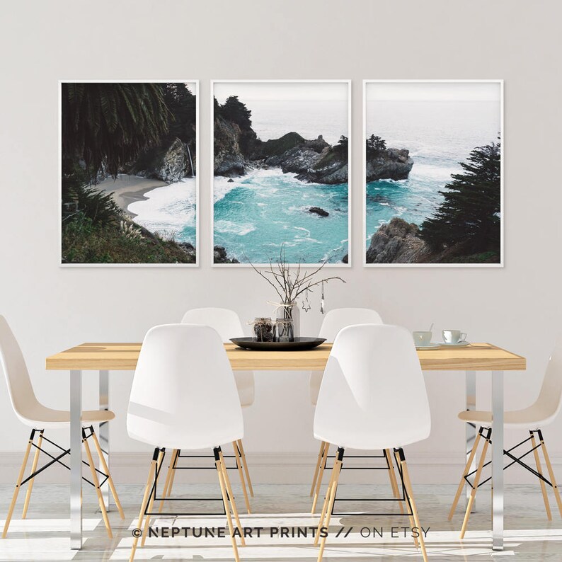 May include: Three framed photographs of a rocky coastline with turquoise water. The photos are hung on a white wall above a wooden table with white chairs.