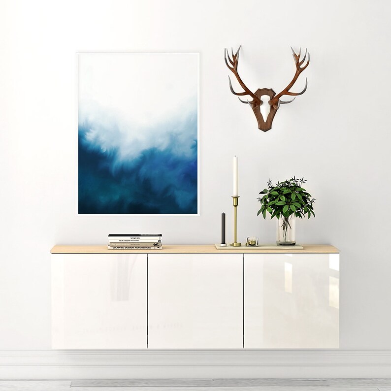 May include: White cabinet with three doors, a wooden top, and a blue and white abstract painting hanging on the wall above it. A set of books and a small plant are on the top of the cabinet. A set of antlers is mounted on the wall to the right of the painting.