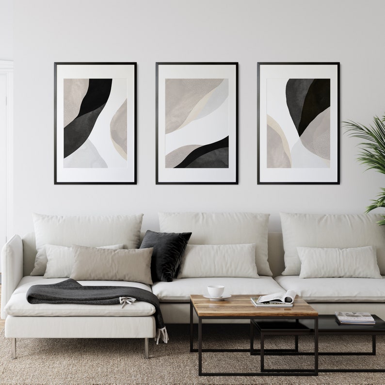 May include: Three framed abstract art prints with black and white shapes on a beige background. The prints are hanging on a white wall above a white sectional sofa with black and beige throw pillows.