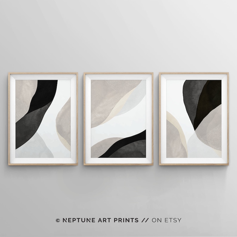 May include: Set of three abstract art prints in a minimalist style. The prints feature a combination of black, white, and beige colors in a fluid, organic design. The prints are framed in a light wood frame.