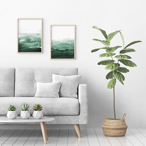 May include: A white sofa with two throw pillows and three small potted succulents sits in front of a white wall with two framed watercolour paintings of green landscapes. A large potted plant with green leaves stands to the right of the sofa.