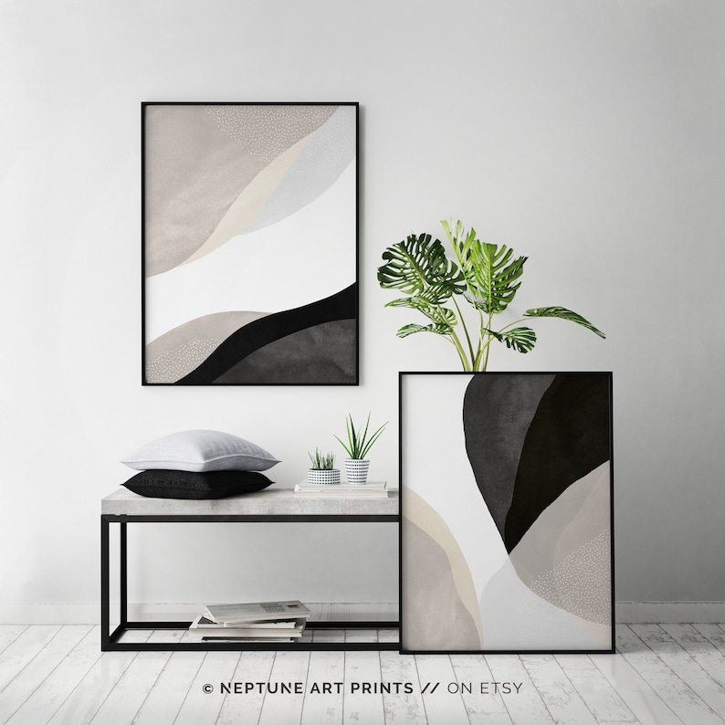 May include: Two abstract art prints in black frames with black, white, and beige shapes. The prints are hanging on a white wall above a concrete shelf with a potted plant and two throw pillows.