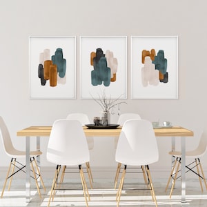 May include: Three abstract art prints with a white background and colorful brushstrokes in shades of teal, brown, and beige. The prints are framed in white and hung on a white wall above a wooden dining table with white chairs.