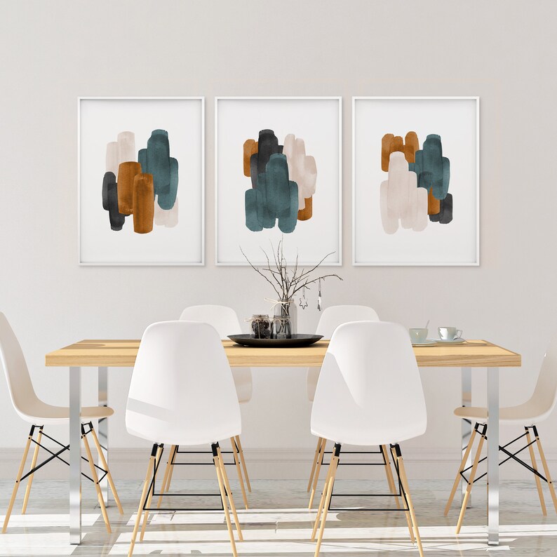 May include: Three abstract art prints with a white background and colorful brushstrokes in shades of teal, brown, and beige. The prints are framed in white and hung on a white wall above a wooden dining table with white chairs.