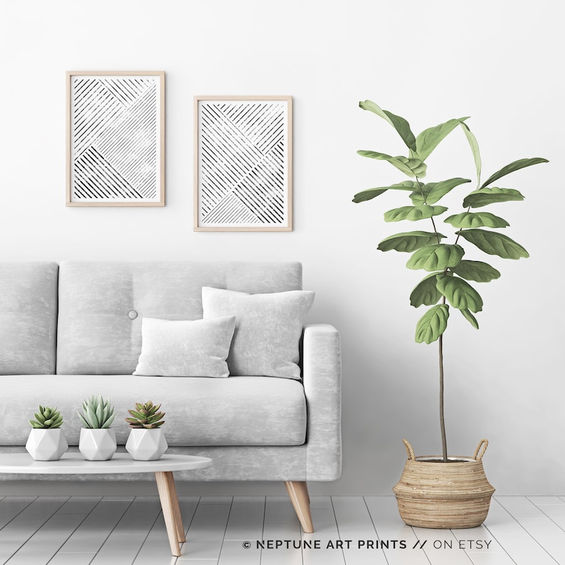May include: Two black and white abstract geometric prints in wooden frames hang on a white wall above a gray sofa with a green plant in a woven basket.