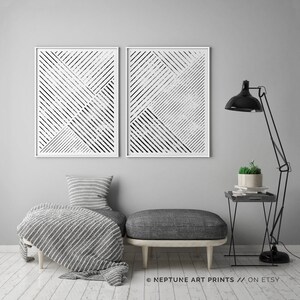 May include: Two black and white abstract geometric prints with a white frame. The prints are hanging on a gray wall above a gray couch with a white throw blanket. A black lamp with a black shade is on the right side of the image. A small black side table with a potted plant is next to the lamp.