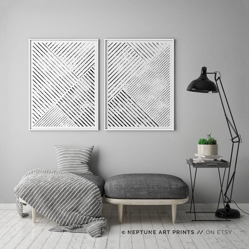 May include: Two black and white abstract geometric prints with a white frame. The prints are hanging on a gray wall above a gray couch with a white throw blanket. A black lamp with a black shade is on the right side of the image. A small black side table with a potted plant is next to the lamp.