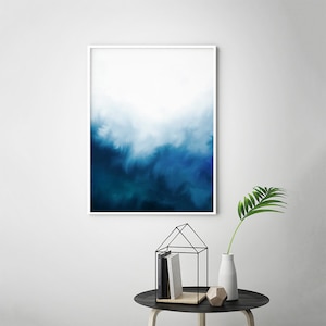 May include: Abstract art print with a white frame. The print features a gradient of blue shades, transitioning from a light blue at the top to a dark blue at the bottom. The blue shades are reminiscent of a cloudy sky or ocean waves.