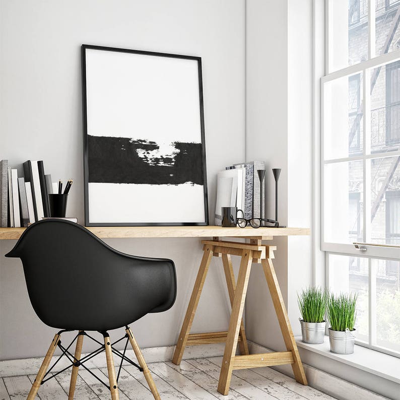 May include: A black and white abstract art print in a black frame hangs above a wooden desk with a black chair. The desk is supported by two wooden sawhorses. There are two potted plants on the floor next to the desk.