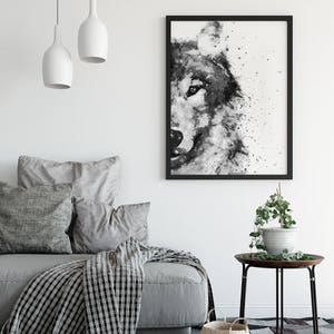 May include: Black and white watercolour painting of a wolf's face with a splatter effect. The painting is framed in a black frame and hanging on a white wall.