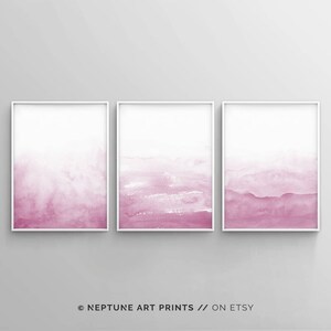 May include: Set of three abstract watercolor prints in shades of pink. The prints are framed in white and feature a gradient of pink from light to dark.