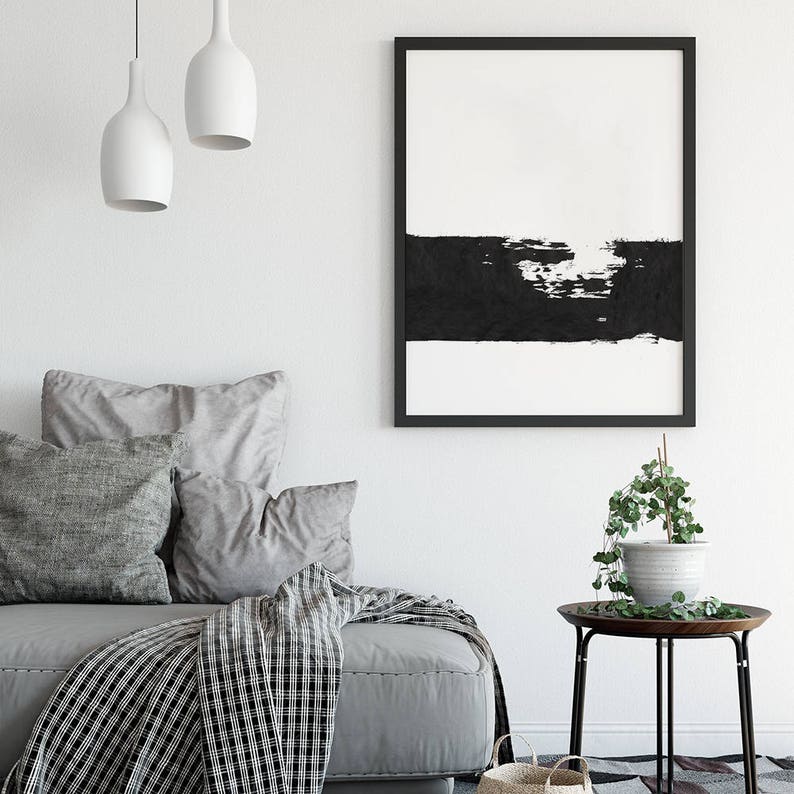 May include: Black and white abstract art print in a black frame hanging on a white wall above a gray couch with pillows and a black and white throw blanket. A small side table with a potted plant sits in the corner.