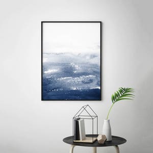 May include: A framed abstract watercolour painting with shades of blue and white. The painting depicts a landscape with a horizon line and a sky with clouds. The painting is framed in a black frame.