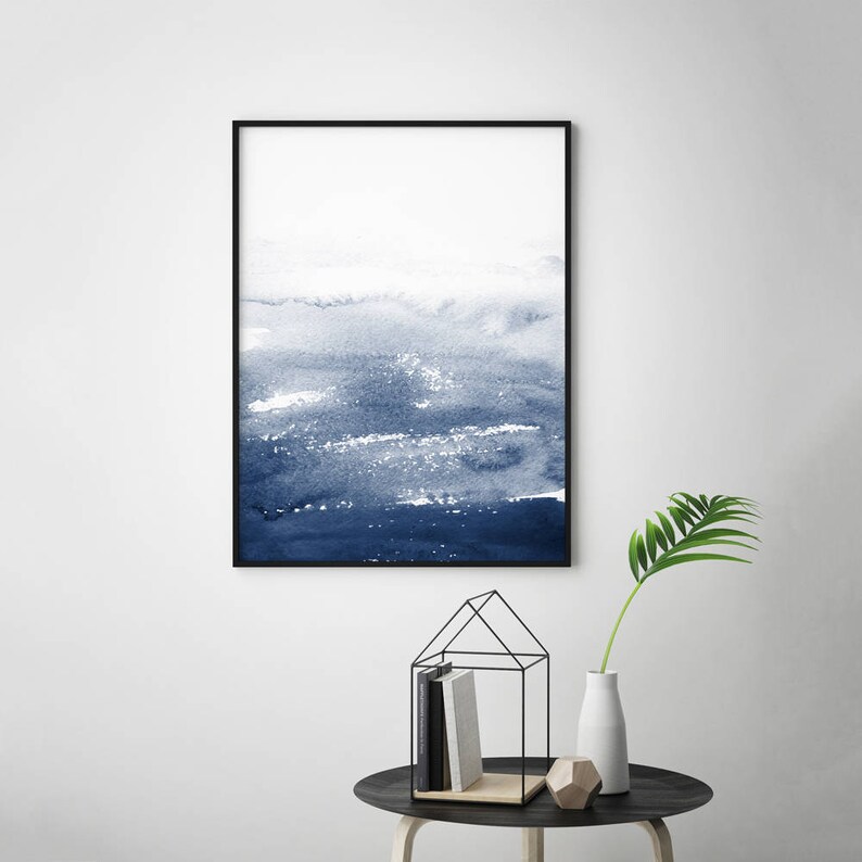 May include: A framed abstract watercolour painting with shades of blue and white. The painting depicts a landscape with a horizon line and a sky with clouds. The painting is framed in a black frame.