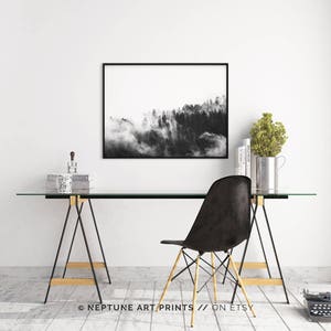 May include: Black and white photograph of a foggy forest landscape. The photo is framed in a black frame and hanging on a white wall above a glass desk with a black chair.