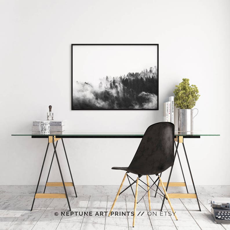 May include: Black and white photograph of a foggy forest landscape. The photo is framed in a black frame and hanging on a white wall above a glass desk with a black chair.