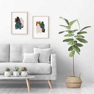 May include: Two framed abstract paintings with muted green, brown, and white brushstrokes hang on a white wall above a gray sofa with white pillows. A potted plant with green leaves sits on the floor next to the sofa.