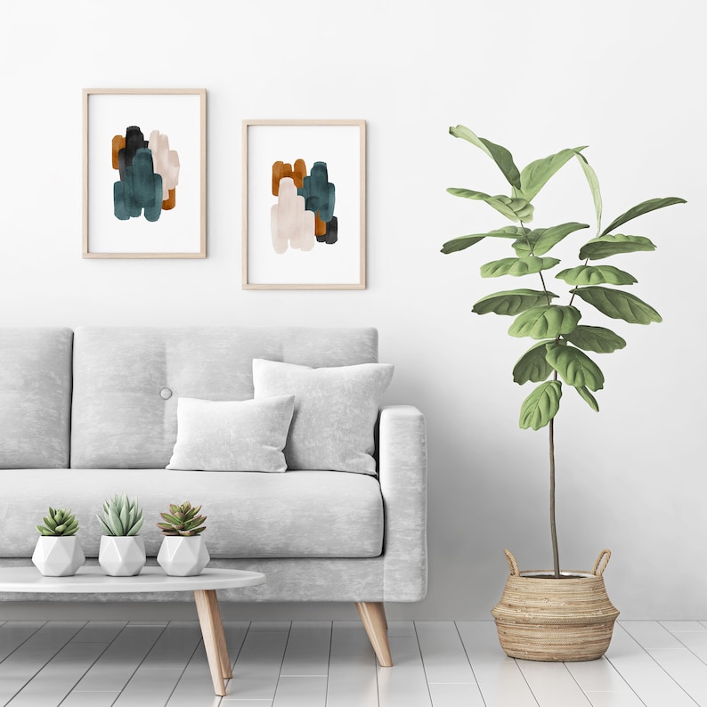 May include: Two framed abstract paintings with muted green, brown, and white brushstrokes hang on a white wall above a gray sofa with white pillows. A potted plant with green leaves sits on the floor next to the sofa.