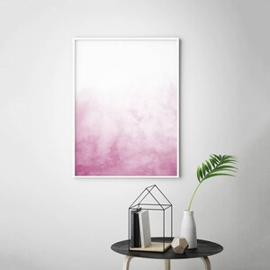 May include: A framed abstract watercolor painting with a pink and white ombre effect. The painting is in a white frame and is hanging on a white wall.