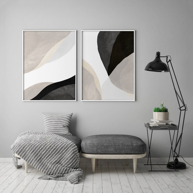 May include: Two abstract art prints in white frames with black and beige shapes on a white background. The prints are hanging on a gray wall above a gray ottoman with a white and gray striped throw blanket.