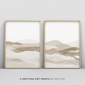 May include: Two framed abstract watercolor prints with a minimalist design featuring brown and beige layered mountains. The prints are in a light wood frame and are hung on a white wall.
