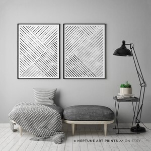 May include: Two black and white abstract geometric prints with a striped pattern. The prints are framed and hung on a gray wall above a gray ottoman and a black side table with a plant on top.