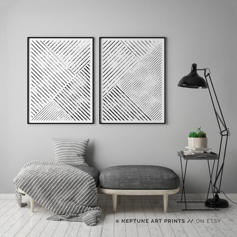 May include: Two black and white abstract geometric prints with a striped pattern. The prints are framed and hung on a gray wall above a gray ottoman and a black side table with a plant on top.