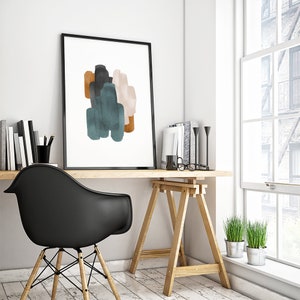 May include: A framed abstract print with a white background and colorful brushstrokes in shades of teal, brown, and pink. The print is hanging on a white wall above a wooden desk with a black chair and a wooden easel.