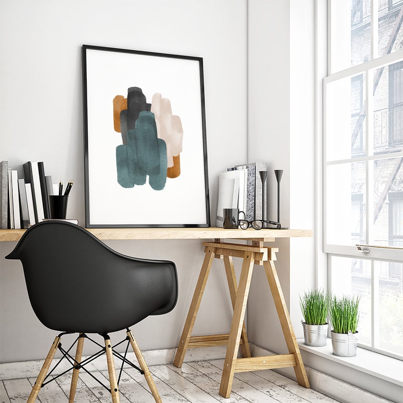 May include: A framed abstract print with a white background and colorful brushstrokes in shades of teal, brown, and pink. The print is hanging on a white wall above a wooden desk with a black chair and a wooden easel.