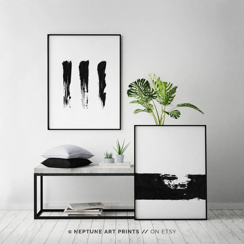 May include: Two black and white abstract art prints with black brushstrokes on a white background. The prints are framed in black and are hanging on a white wall above a concrete shelf with a potted plant and two pillows.